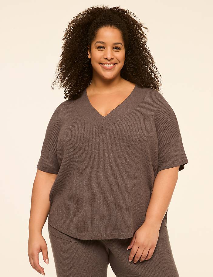 A smiling woman with curly hair wears a v-neck sweater and pants against a plain, light-colored background.