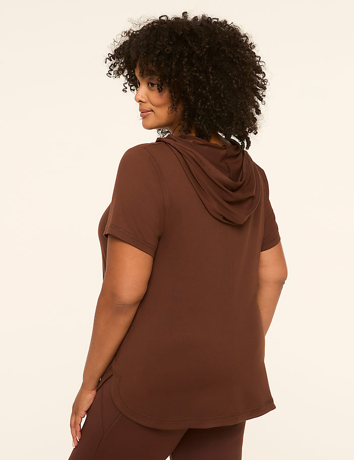 A woman with curly dark hair wears a brown short-sleeved hoodie and leggings, standing against a plain cream background.