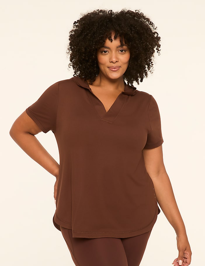 A woman with dark, curly hair and a warm smile poses in a brown polo shirt and leggings against a plain, light-colored background.