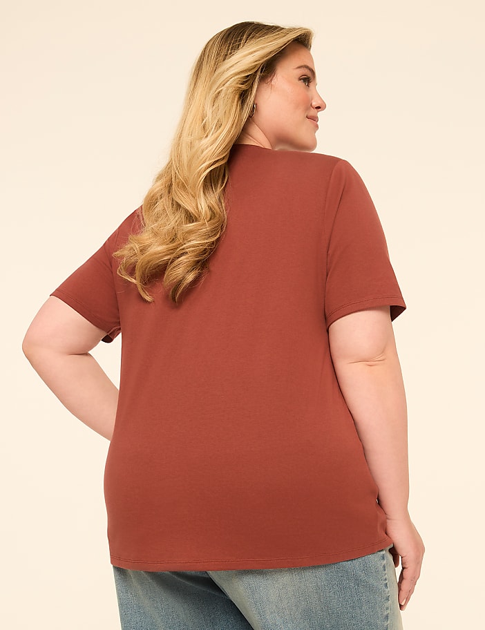 A woman with blonde hair wearing a rust-colored t-shirt and blue jeans stands against a plain, light-colored background.