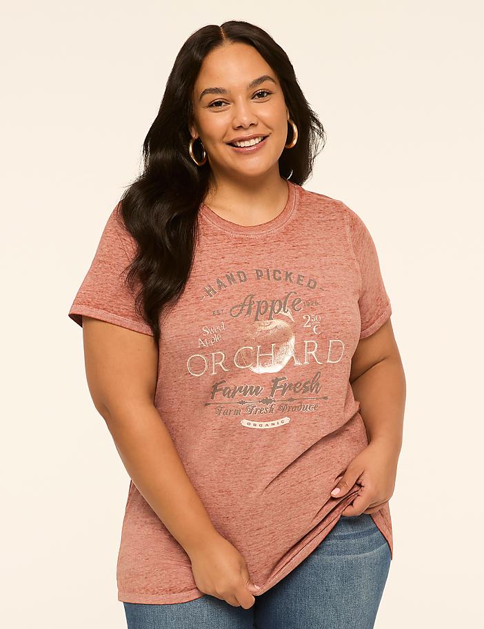 A woman in a pink t-shirt with an apple orchard graphic stands against a plain, light-colored background.