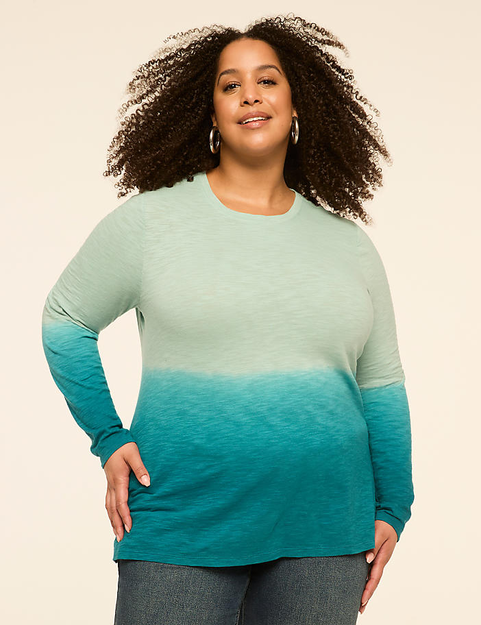 A woman with curly hair smiles at the camera, wearing a long-sleeved, ombre teal shirt and jeans against a plain, light-colored background.