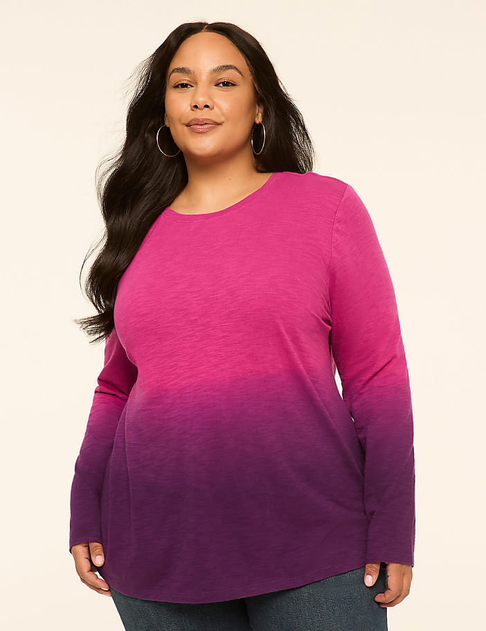 A woman with long dark hair and hoop earrings poses in a long-sleeved, ombre pink and purple shirt against a plain, light-colored background.