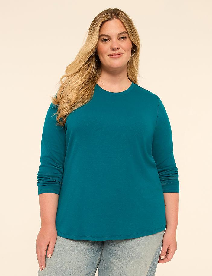 A woman with blonde hair and a teal long-sleeved shirt stands against a plain, light-colored background.