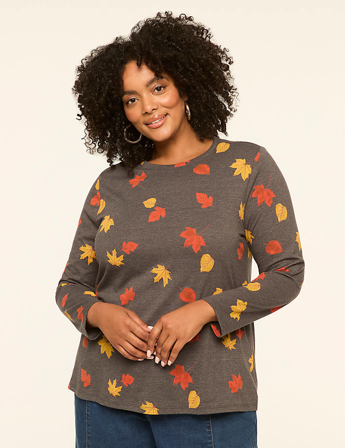 A woman with curly dark hair smiles at the camera, wearing a long-sleeved gray shirt with a pattern of red and yellow leaves, against a plain cream-colored background.