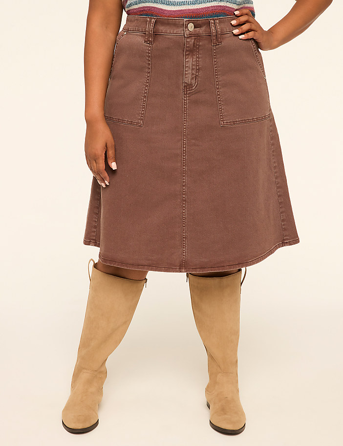 A woman in a brown denim skirt and tan boots stands against a plain cream background.