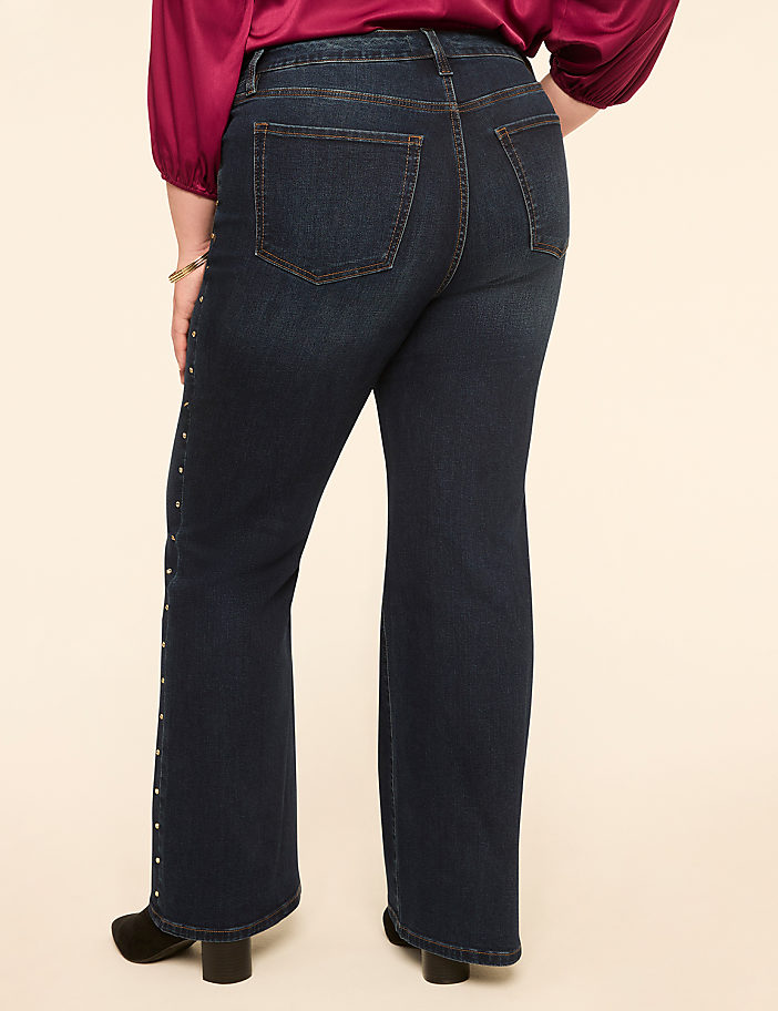 A person wearing dark wash flared jeans with stud detailing down the side stands against a plain, light-colored background.
