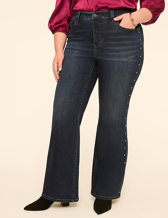 A woman in dark wash flared jeans with stud detailing stands against a plain, light-colored background.