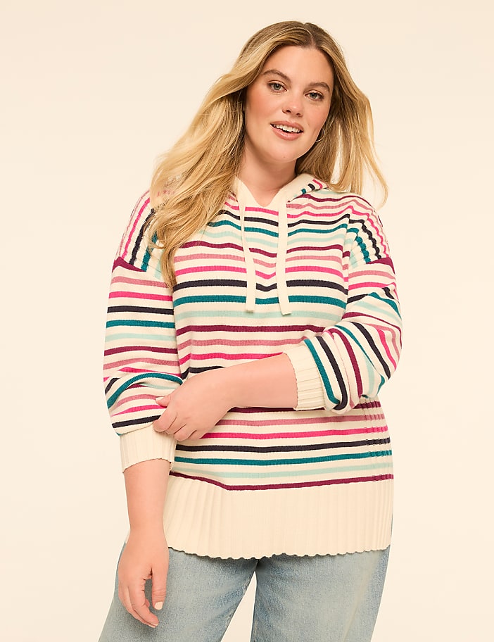 A woman with blonde hair smiles at the camera, wearing a striped hoodie and jeans against a plain, light-colored background.