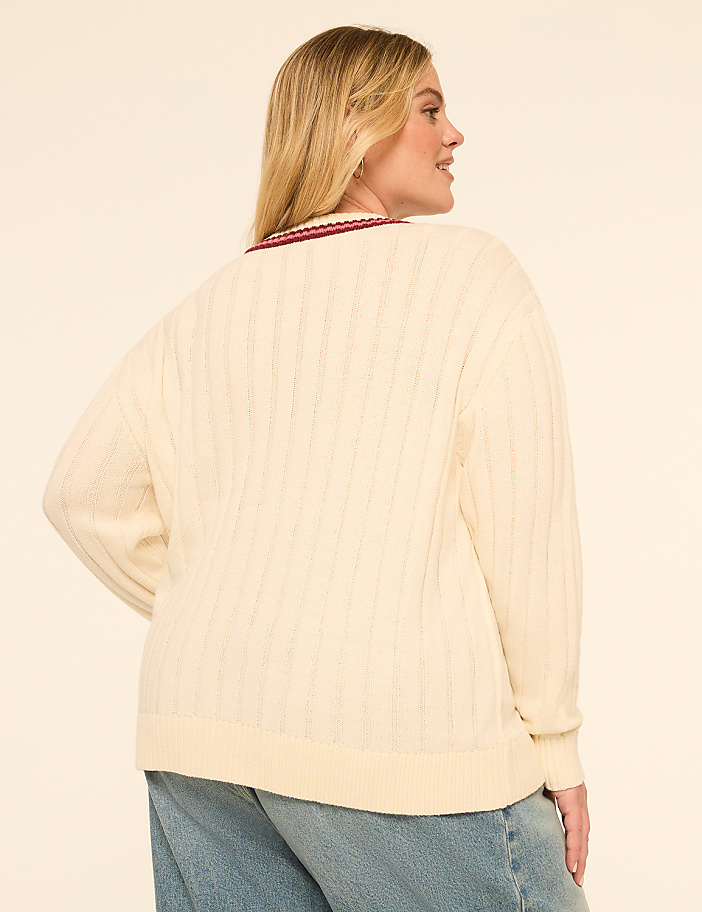 A woman in a cream cable-knit sweater and blue jeans stands against a plain, light-colored background.