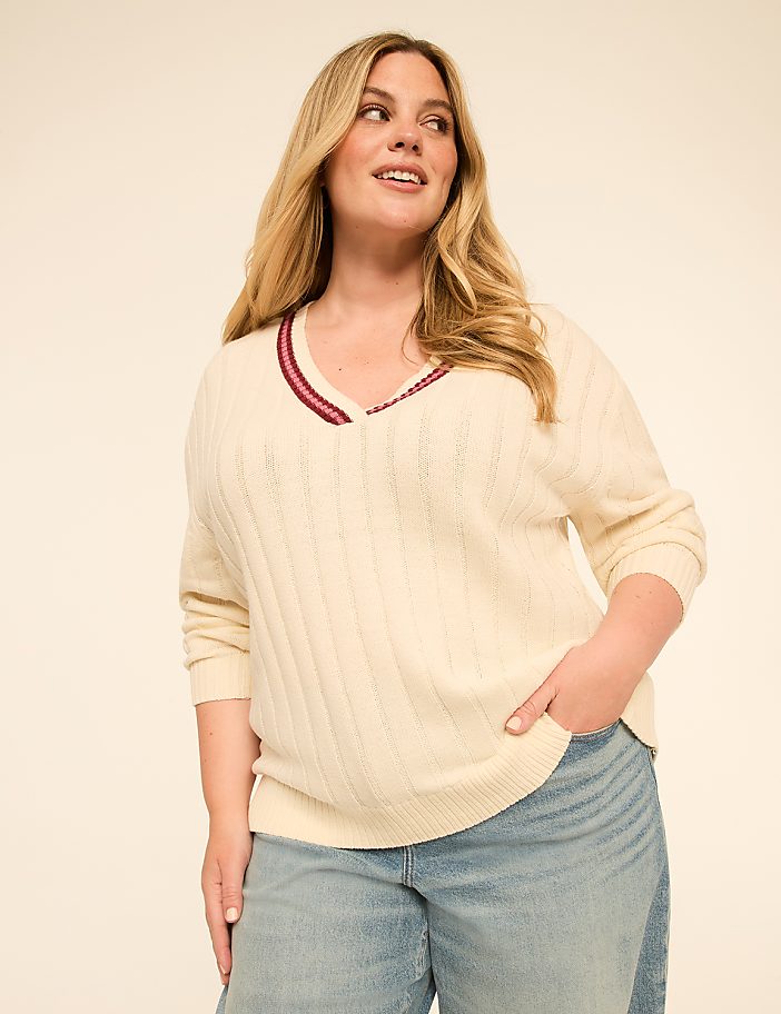 A woman in a cream-colored v-neck sweater and light-wash jeans stands against a plain, light-colored background.