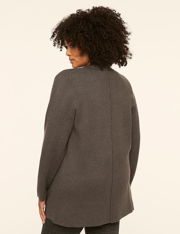 A woman with dark, curly hair stands facing away from the camera in a charcoal gray cardigan and patterned pants against a plain, light background.