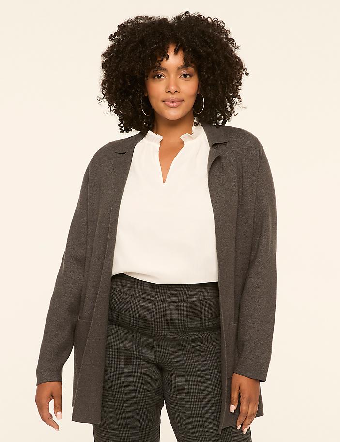 A woman with curly dark hair stands in the foreground wearing a gray cardigan over a white blouse and plaid pants, against a plain, light-colored background.