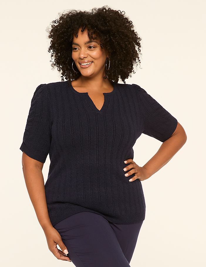 A smiling woman with curly hair poses in a navy blue sweater and pants against a plain, light-colored background.