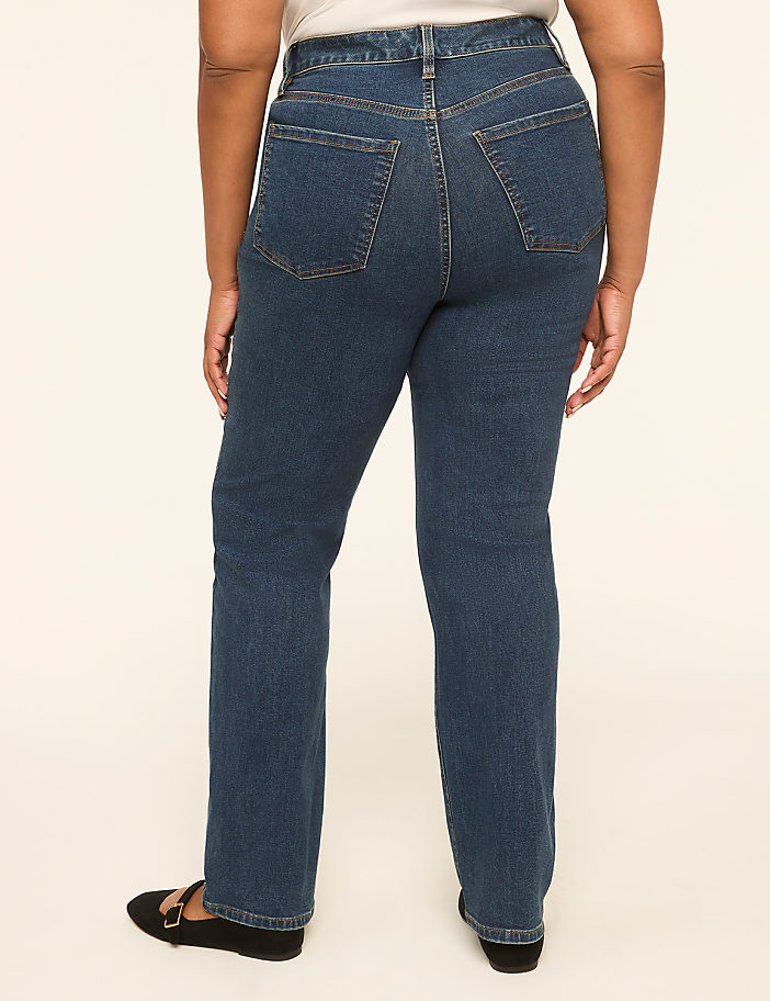 A person wearing dark wash denim jeans and black flats stands against a plain, light-colored background.