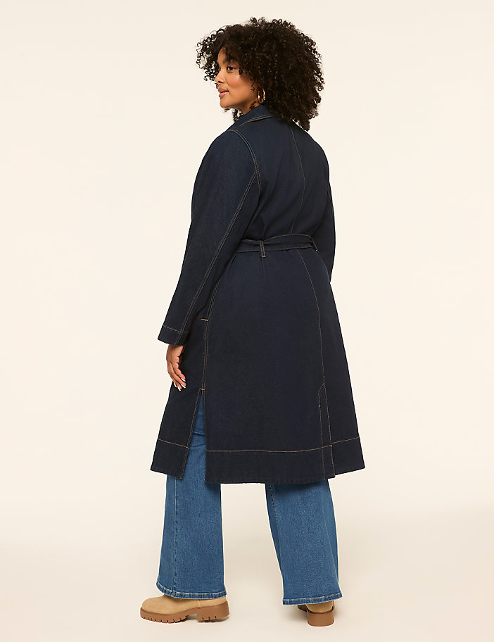 A woman in a denim trench coat and wide-leg jeans stands against a plain, light-colored background.