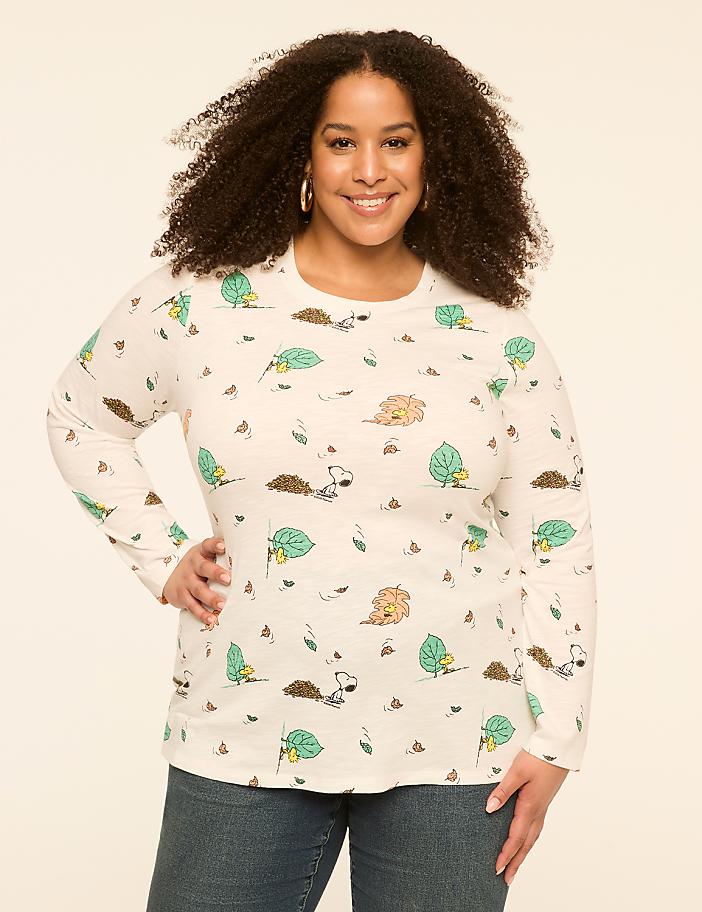 A smiling woman with curly hair poses in a cream-colored long-sleeved shirt with a repeating pattern of cartoon hedgehogs and leaves against a plain, light-colored background.