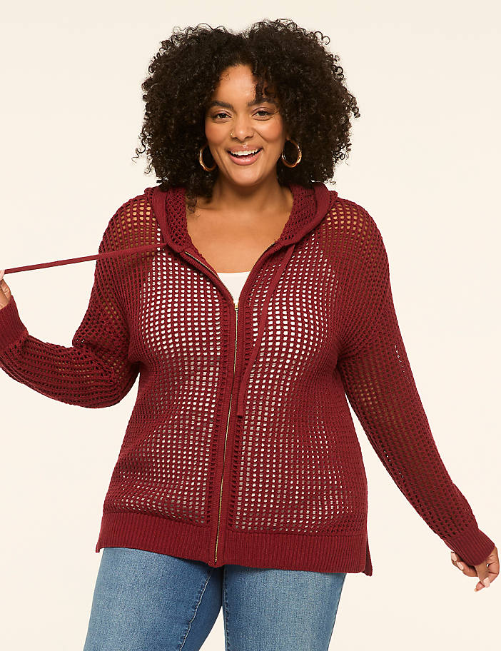 A smiling woman in a maroon open-knit zip-up hoodie stands against a plain cream background.