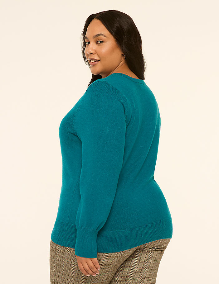 A woman in a teal sweater and plaid pants stands against a plain, light-colored background.