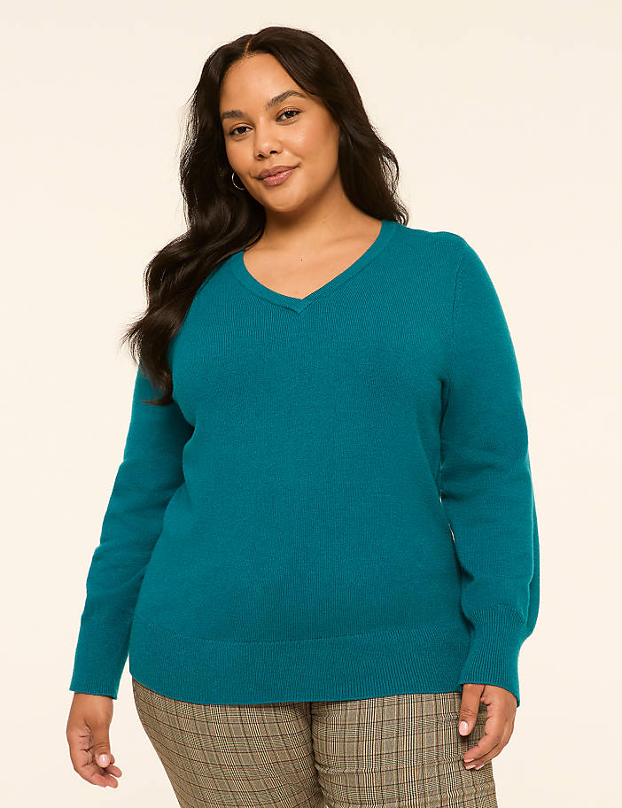 A woman in a teal v-neck sweater and plaid pants stands against a plain, light-colored background.