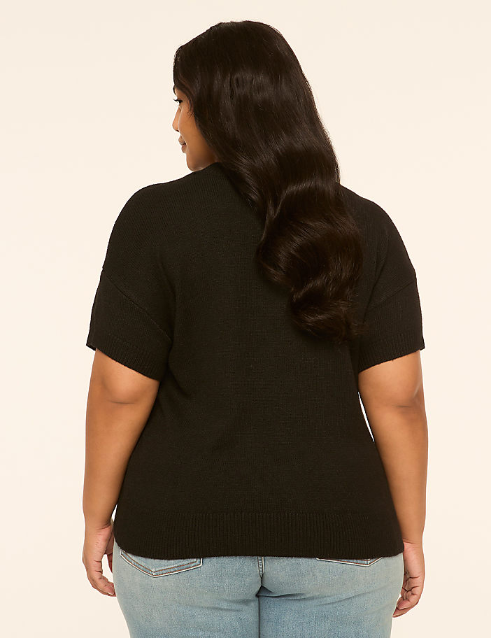 A woman in a black short-sleeved sweater and blue jeans stands against a plain, light-colored background.