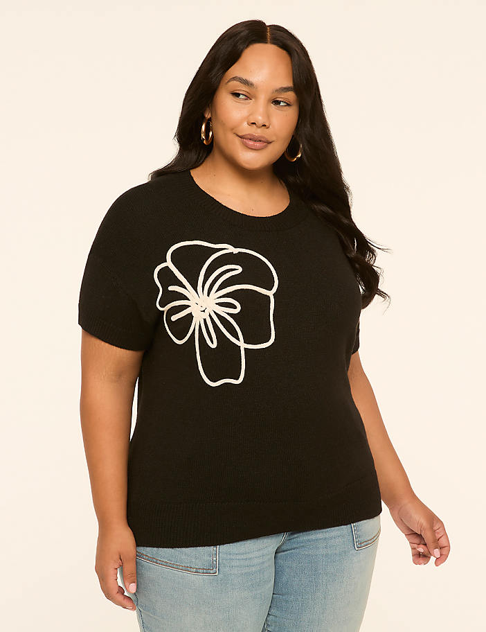 A woman in a black sweater with a white floral design stands against a plain, light-colored background.