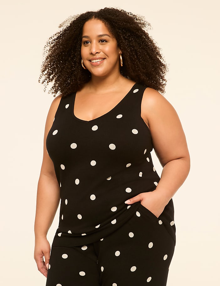 A smiling woman with curly hair in a black and white polka dot tank top stands against a plain, light-colored background.