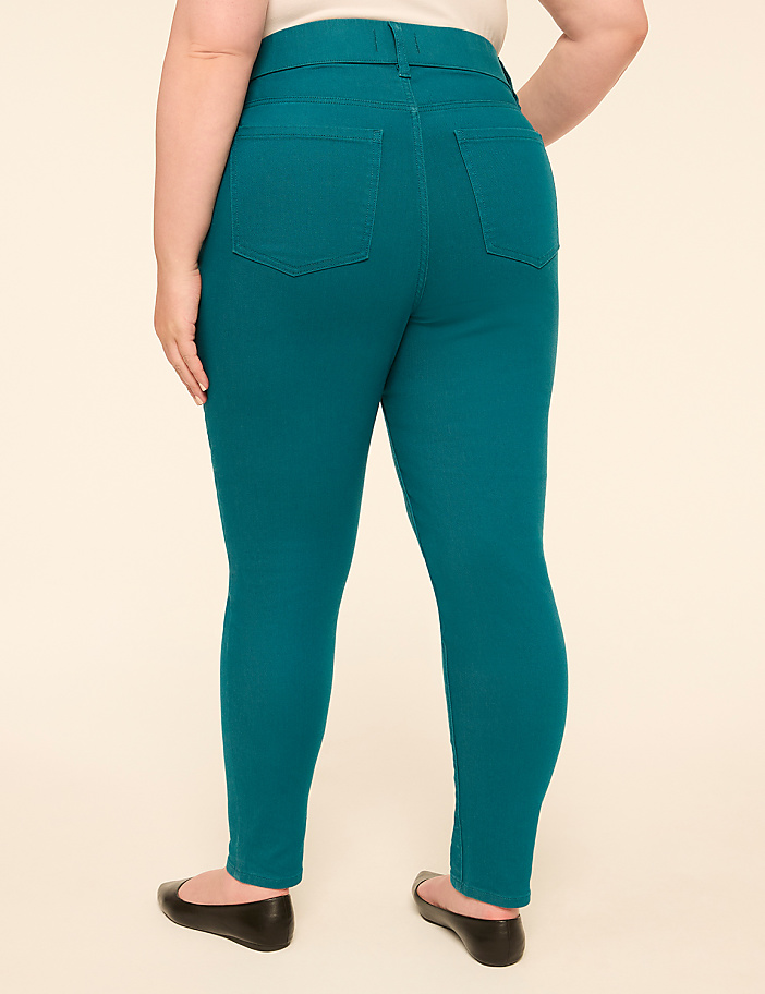 A person wearing teal skinny jeans and black flats stands against a plain, light-colored background.