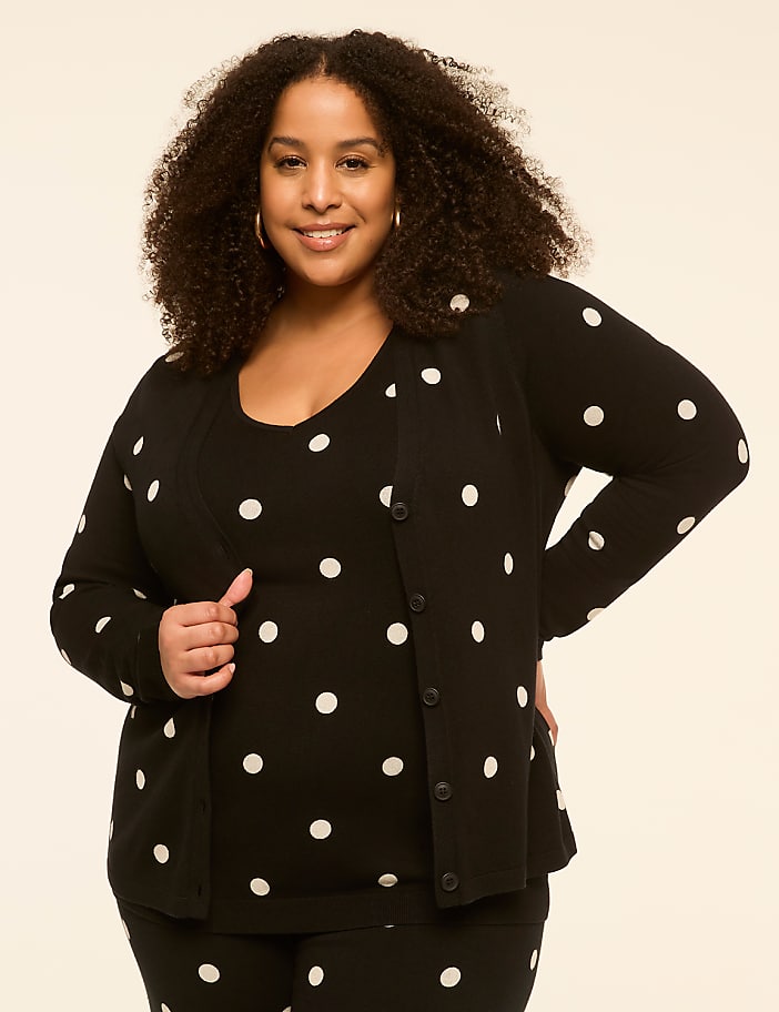 A smiling woman with curly hair poses in a black and white polka dot outfit against a plain, light-colored background.