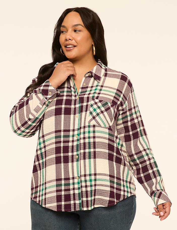 A woman in a cream, purple, and green plaid shirt stands against a plain, light-colored background.