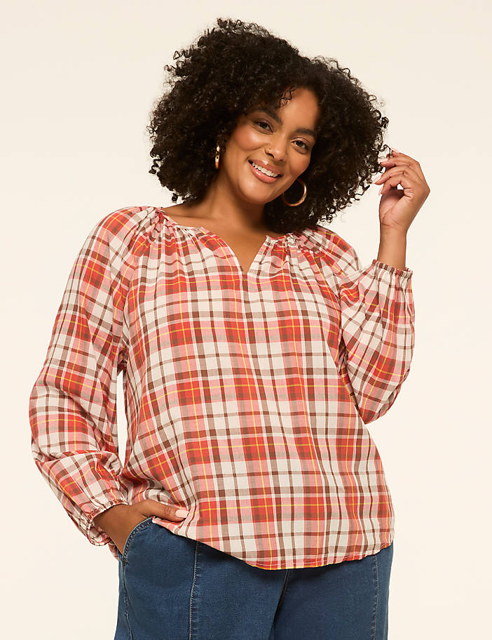 A smiling woman with curly hair wears a plaid blouse and jeans against a plain background.