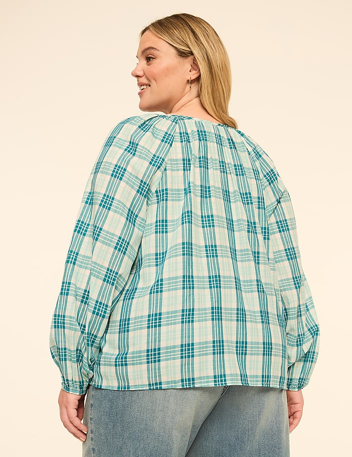 A woman in a teal plaid blouse and denim jeans stands against a plain, light-colored background.