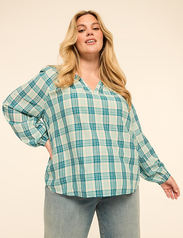 A woman in a teal and white plaid blouse and light blue jeans poses against a plain, light beige background.