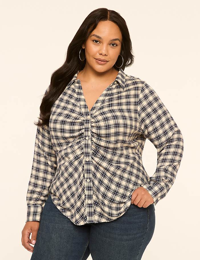 A woman in a plaid shirt and jeans stands against a plain, light-colored background.