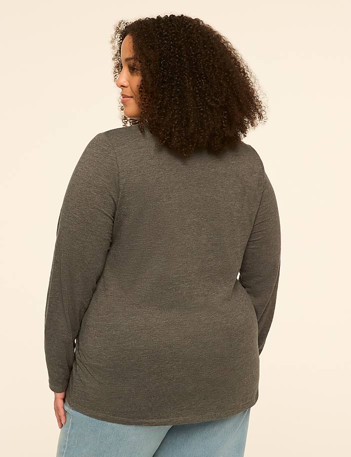 A woman with curly hair, seen from the back, wears a dark gray long-sleeved shirt and light blue jeans against a plain, light-colored background.