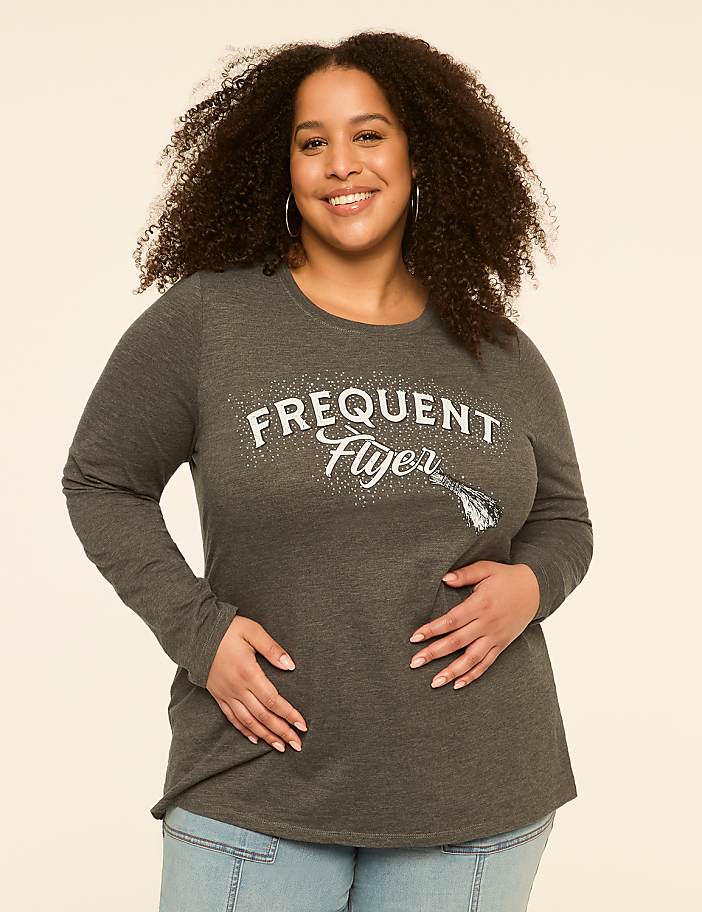 A smiling woman with curly hair in a grey long-sleeved shirt with "Frequent Flyer" printed on it stands against a plain, light-colored background.