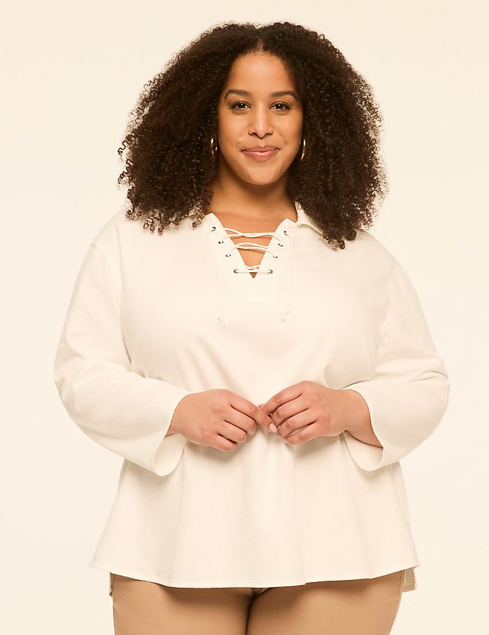 A woman with curly dark hair and a white lace-up shirt stands against a plain, light-colored background.