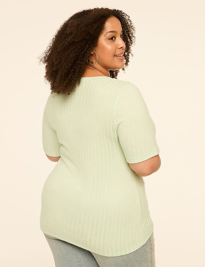 A woman with curly hair in a light green ribbed shirt stands against a plain, light-colored background.