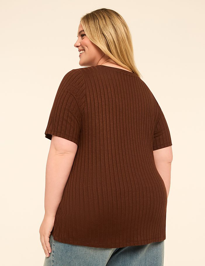 A woman in a brown ribbed t-shirt and jeans smiles over her shoulder against a plain, light-colored background.