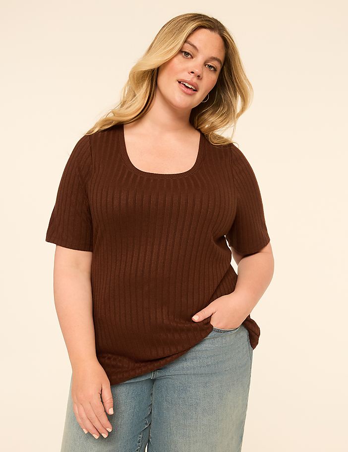 A woman with blonde hair and fair skin, wearing a brown ribbed t-shirt and light blue jeans, stands against a plain, light beige background.