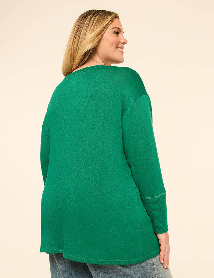 A woman in a green sweater and jeans stands against a plain, light-colored background.