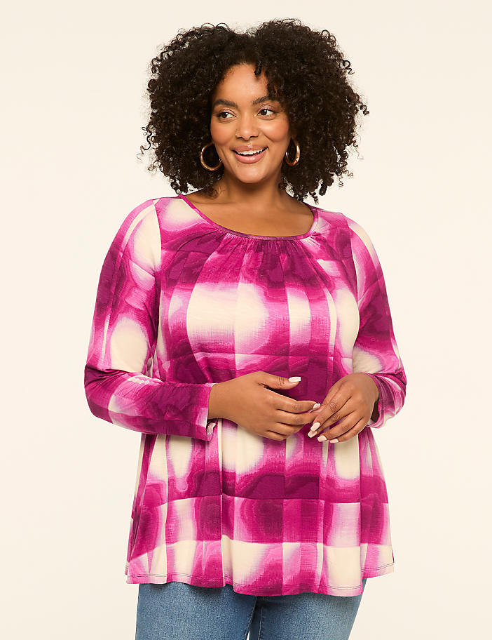A smiling woman with curly dark hair and hoop earrings poses in a pink and cream patterned top and blue jeans against a plain cream background.