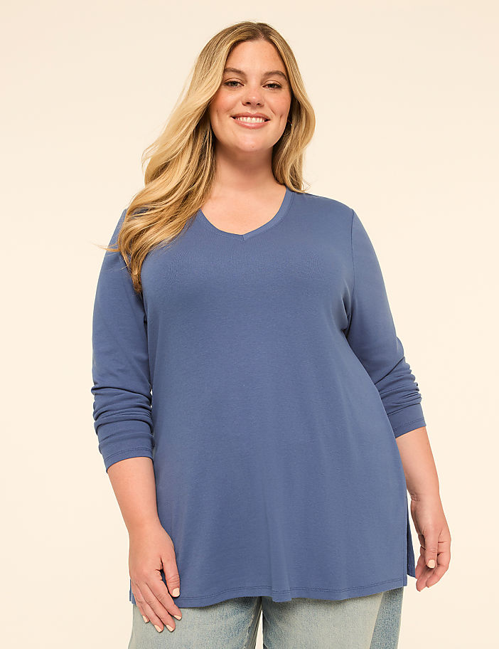 A smiling woman with blonde hair wears a blue long-sleeved shirt and jeans against a plain, light-colored background.