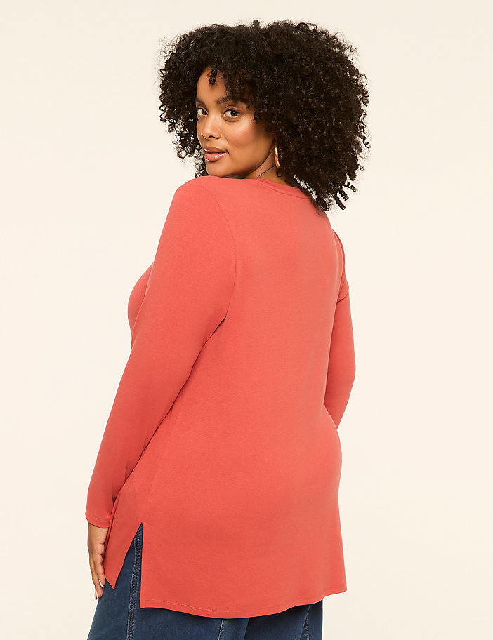A woman in a coral-colored tunic and blue jeans stands against a plain, light-colored background.