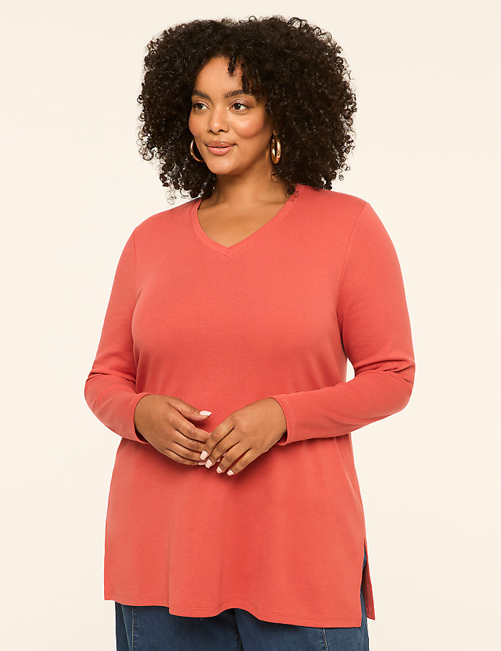 A woman with curly dark hair and hoop earrings poses in a coral v-neck shirt against a plain cream background.