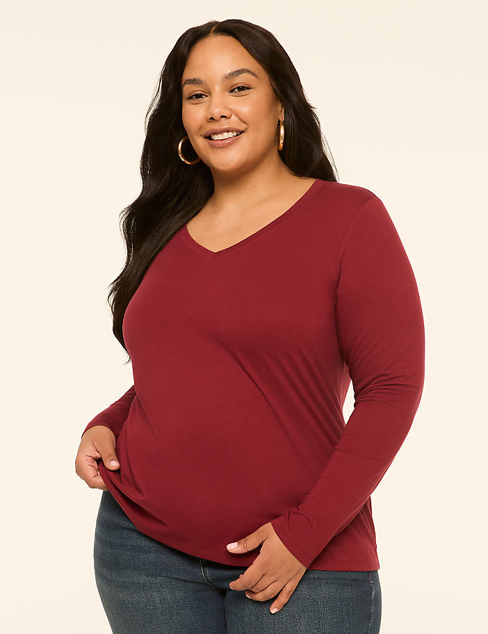 A smiling woman in a maroon v-neck shirt and blue jeans stands against a plain, light-colored background.