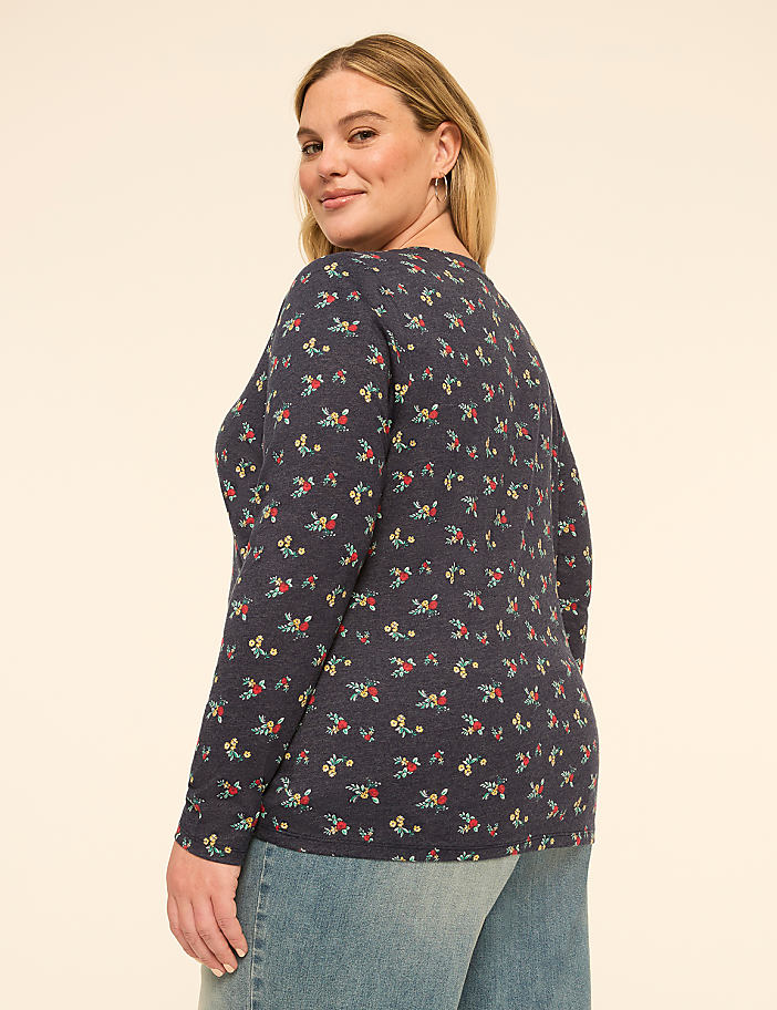 A woman in a floral long-sleeved shirt and jeans stands against a plain, light-colored background.