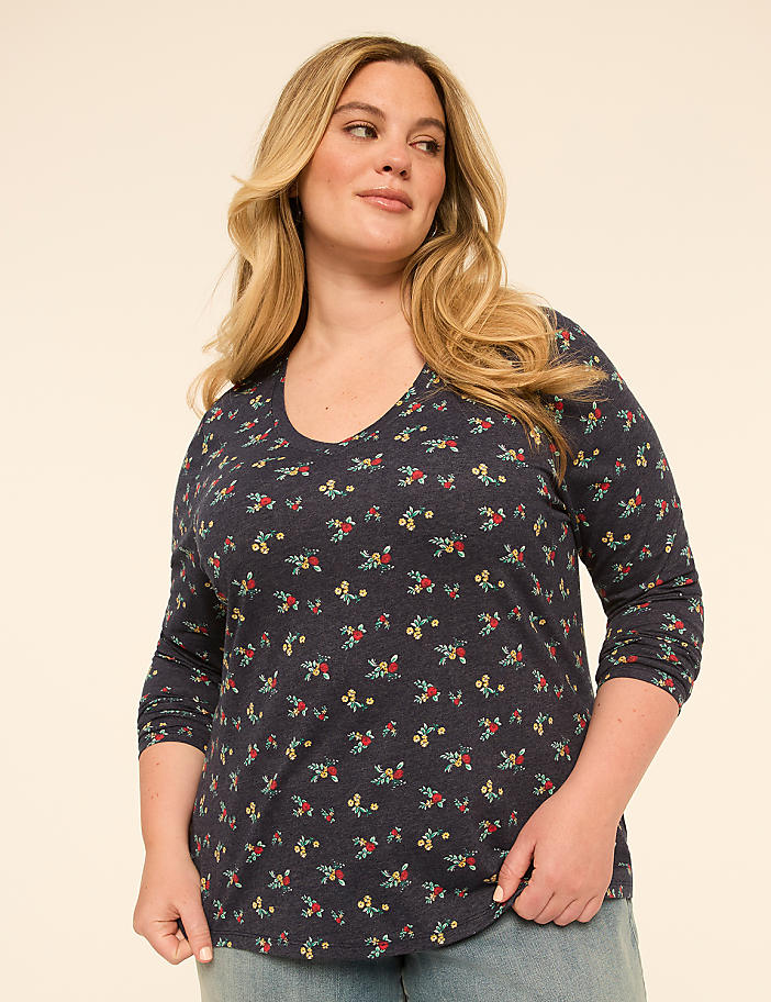 A woman with blonde hair and a floral shirt stands against a plain, light-colored background.