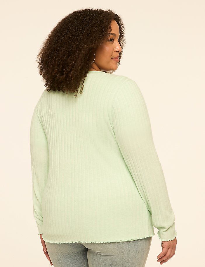 A woman with curly brown hair, wearing a light green ribbed long-sleeved shirt and jeans, stands against a plain, light beige background.