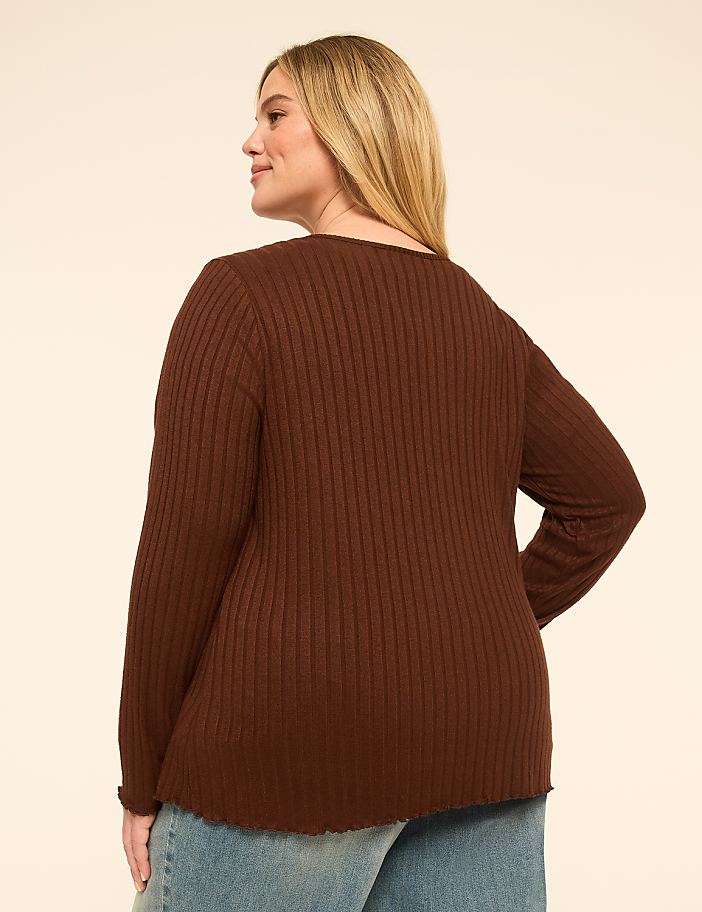 A woman in a brown ribbed sweater and blue jeans stands against a plain, light-colored background.
